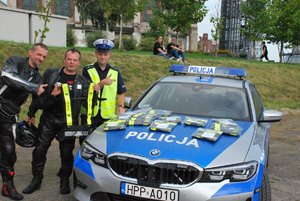 Zdjęcie kolorowe. Widoczny umundurowany policjant ruchu drogowego oraz uczestniczy wydarzenia oraz radiowóz oznakowany