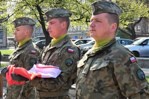 obok siebie stoją trzy osoby żołnierze, jeden z nich trzyma flagę