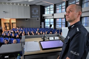 Umundurowany policjant ogląda spot, który wyświetlany jest na ścianie w auli. W tle uczniowie, którzy również patrzą w tym samym kierunku, co policjant.