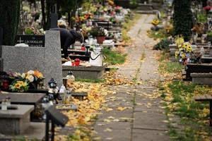 zdjęcie przedstawia zbliżenie na torębkę leżącą na pomniku, obok kobieta zajęta sprzątaniem nagrobka