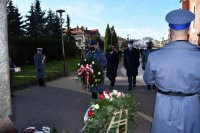 Policjant niosący wieniec, w tle delegacja służb mundurowych