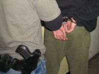 Zdjęcie przedstawia policjanta oraz osobę zatrzymaną w kajdankach.