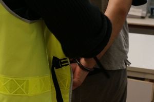 Zdjęcie przedstawia umundurowanego policjanta stojącego za zatrzymanym mężczyzną w kajdankach założonych na ręce z tyłu.