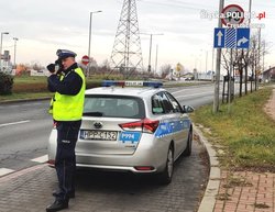 Zdjęcie przedstawia umundurowanego policjanta na tle radiowozu.