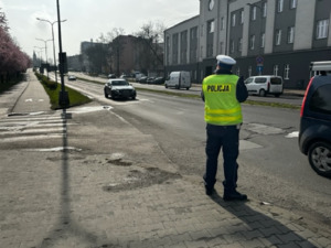 Zdjęcie przedstawia umundurowanego policjanta Wydziału Ruchu Drogowego mierzącego prędkość pojazdów poruszających się po ulicy.