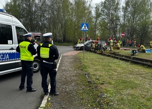 Zdjęcie przedstawia umundurowanych policjantów stojących obok radiowozu. W tle strażacy.