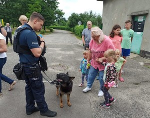 Zdjęcie przedstawia uczestników festynu stojących wokół umundurowanego policjanta z psem służbowym.