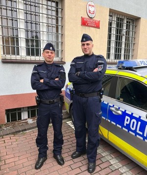 Zdjęcie przedstawia dwóch umundurowanych policjantów stojących przy radiowozie. W tle Komisariat Policji III w Sosnowcu.