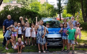 Zdjęcie przedstawia grupę dzieci oraz umundurowanego policjanta. W tle radiowóz służbowy.