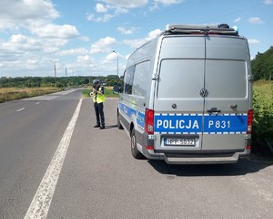 Zdjęcie przedstawia umundurowanego policjanta stojącego obok oznakowanego radiowozu.