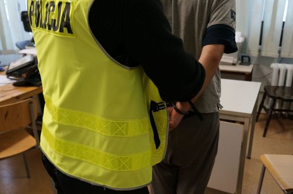 Zdjęcie przedstawia mężczyznę w kajdankach założonych na ręce trzymane z tyłu i policjanta w kamizelce z napisem &quot;Policja&quot;.