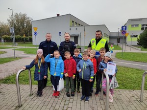 Zdjęcie przedstawia grupę dzieci. Za nimi stoją umundurowani policjanci.