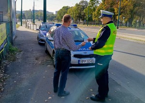 Zdjęcie przedstawia umundurowanego policjanta wręczającego element odblaskowy mężczyźnie. W tle oznakowane radiowozy służbowe.