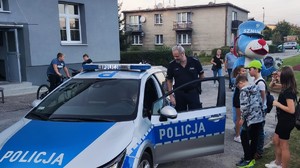 Zdjęcie przedstawia umundurowanego policjanta z grupą dzieci przy radiowozie służbowym.