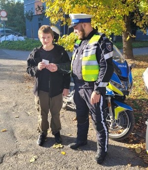 Zdjęcie przedstawia umundurowanego policjanta wręczającego chłopcu element odblaskowy.