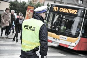 Zdjęcie przedstawia umundurowanego policjanta w kamizelce odblaskowej. W tle autobus.