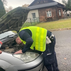 Zdjęcie przedstawia umundurowanego policjanta w kamizelce odblaskowej pochylającego się na samochodem.