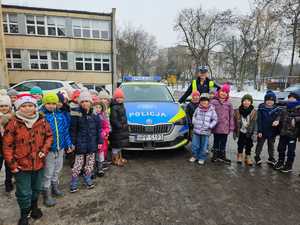 Zdjęcie przedstawia umundurowaną policjantkę z dziećmi. W tle radiowóz.