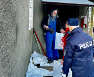 Zdjęcie przedstawia umundurowanych policjantów oraz księdza wręczających przed domem paczkę starszemu mężczyźnie w kapturze.