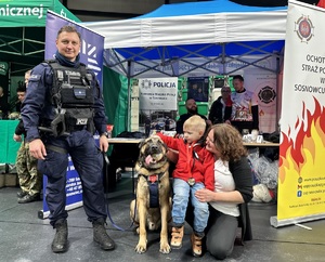 Zdjęcie przedstawia umundurowanego policjanta, psa służbowego oraz uczestników WOŚP.