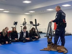 Zdjęcie przedstawia umundurowanego policjanta z psem służbowym. W tle uczniowie.