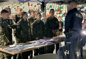 Zdjęcie przedstawia umundurowanego policjanta oraz uczniów stojących przy stoisku profilaktycznym.