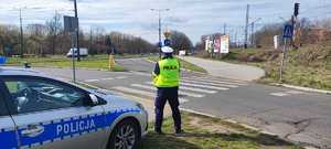 Zdjęcie przedstawia umundurowanego policjanta z wydziału ruchu drogowego przy przejściu dla pieszych. W tle oznakowany radiowóz policyjny.