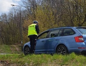 Zdjęcie przedstawia umundurowanego policjanta podczas kontroli drogowej przy niebieskim pojeździe.