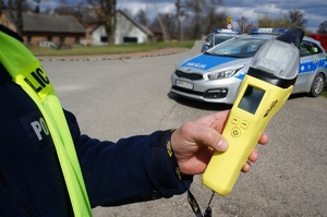 Zdjęcie przedstawia umundurowanego policjanta z alkomatem. W tle oznakowany radiowóz.