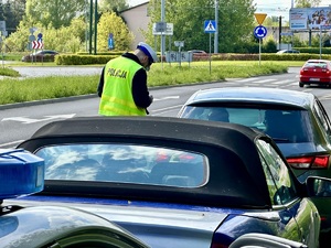 Zdjęcie przedstawia umundurowanego policjanta stojącego przy pojeździe. W tle dwa samochody oraz oznakowany, policyjny radiowóz.