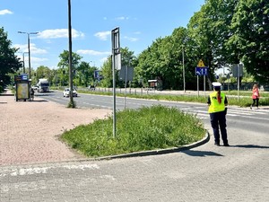Zdjęcie przedstawia umundurowaną policjantkę z Wydziału Ruchu Drogowego w kamizelce odblaskowej, stojącą w rejonie przejścia dla pieszych. Po lewej stronie przystanek autobusowy.