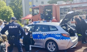 Zdjęcie przedstawia umundurowanego policjanta, strażaków i uczestników festynu. W tle radiowóz policyjny oraz wóz strażacki.