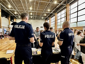 Zdjęcie przedstawia umundurowaną policjantkę oraz dwóch policjantów przy stoisku policyjnym na sali gimnastycznej.