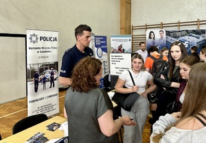 Zdjęcie przedstawia umundurowanego policjanta oraz pracownicę Zespołu Kadr i Szkolenia rozmawiających z grupą uczennic przy stoisku.