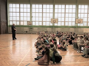 Zdjęcie przedstawia umundurowanego policjanta przemawiającego do dużej grupy dzieci na sali gimnastycznej w szkole.