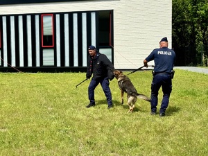 Zdjęcie przedstawia dwóch umundurowanych policjantów i psa służbowego podczas pokazu.