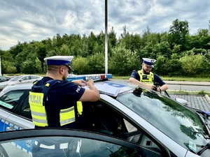 Zdjęcie przedstawia dwóch, umundurowanych policjantów wykonujących czynności przy oznakowanym radiowozie.