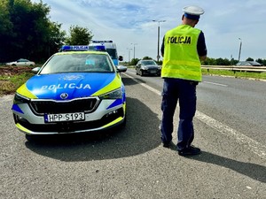 Zdjęcie przedstawia umundurowanego policjanta przy dwóch oznakowanych radiowozach mierzącego prędkość pojazdów.