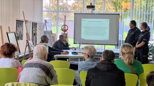 Zdjęcie przedstawia umundurowanych policjantów z grupą seniorów. W tle prezentacja multimedialna.