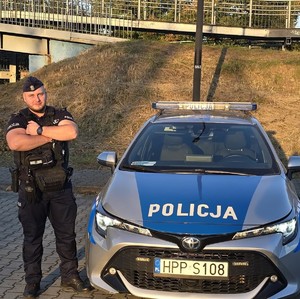 Zdjęcie przedstawia umundurowanego policjanta stojącego obok oznakowanego radiowozu.