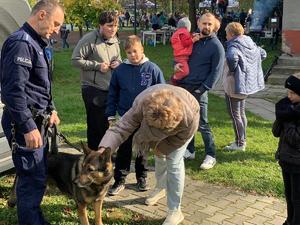 Zdjęcie przedstawia umundurowanego policjanta z psem służbowym oraz uczestnikami podczas festynu.