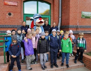 Zdjęcie przedstawia grupę dzieci, policyjną maskotkę, Sznupka oraz umundurowanych policjantów przed budynkiem Komisariatu Policji II w Sosnowcu.