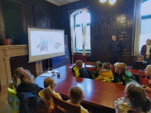 Zdjęcie przedstawia grupę dzieci siedzącą przy stole w Komisariacie Policji II w Sosnowcu. W tle prezentacja multimedialna.