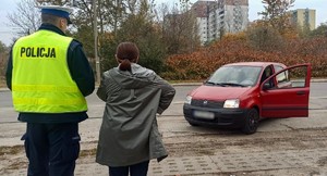 Zdjęcie przedstawia umundurowanego policjanta oraz stojąca obok kobietę. W tle czerwony samochód.