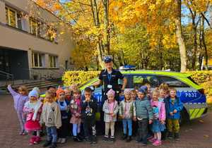 Zdjęcie przedstawia umundurowaną policjantkę z grupą dzieci na tle radiowozu przed przedszkolem.