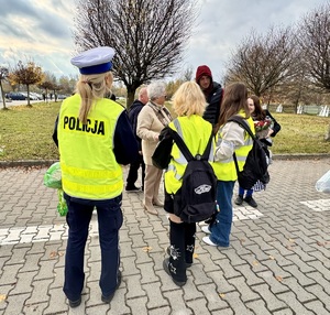 Zdjęcie przedstawia umundurowaną policjantkę i dwie uczennice w kamizelkach odblaskowych wręczające ludziom elementy odblaskowe.