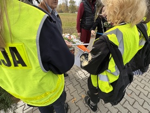 Zdjęcie przedstawia umundurowaną policjantkę i dwie uczennice w kamizelkach odblaskowych wręczające ludziom elementy odblaskowe.