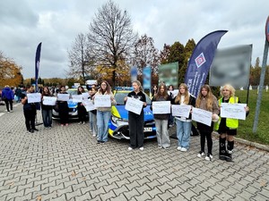 Zdjęcie przedstawia grupę dzieci z kartkami w rękach na tle radiowozów służbowych przy cmentarzu.