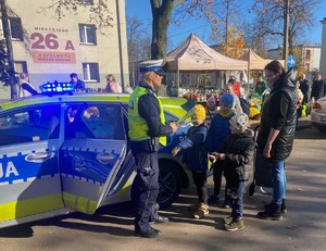 Zdjęcie przedstawia umundurowaną policjantkę z grupą ludzi w rejonie cmentarza. W tle oznakowany radiowóz policyjny.