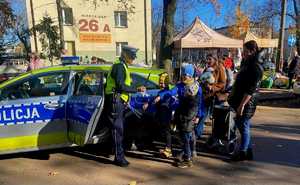 Zdjęcie przedstawia umundurowaną policjantkę z grupą ludzi w rejonie cmentarza. W tle oznakowany radiowóz policyjny.